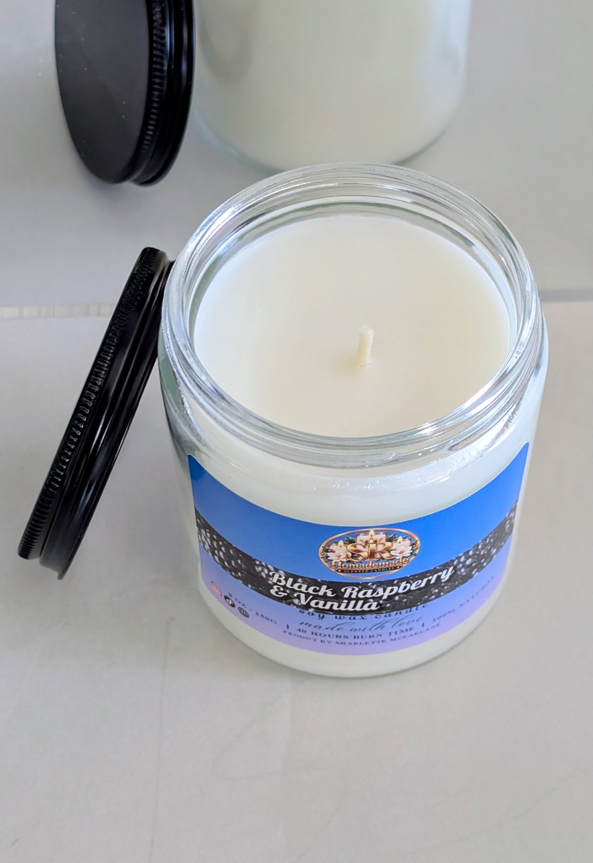 A white wax candle in a clear glass jar with a blue and lavender label reading 'Black Raspberry & Vanilla,' shown with its black lid beside it on a light surface.